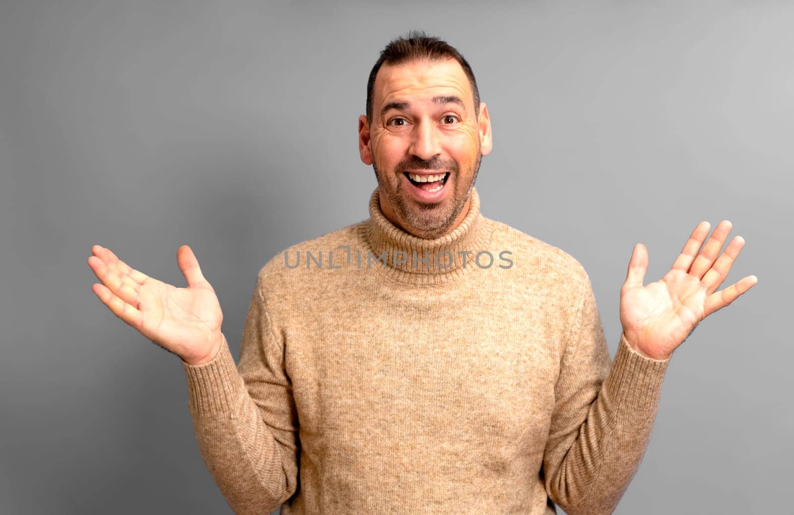 Bearded Hispanic man in his 40s wearing a turtleneck sweater with arms outstretched in an expression of surprise, isolated on gray studio background