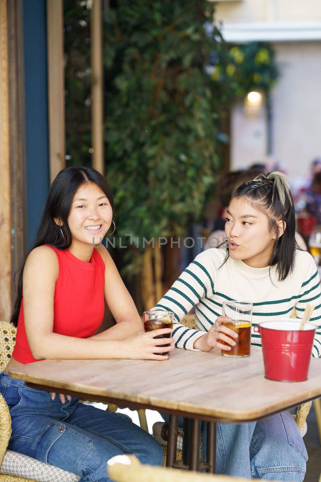 Cheerful young Asian female friends, in casual apparel sitting at table with cold drinks and speaking about plans for future while spending time together in cafe