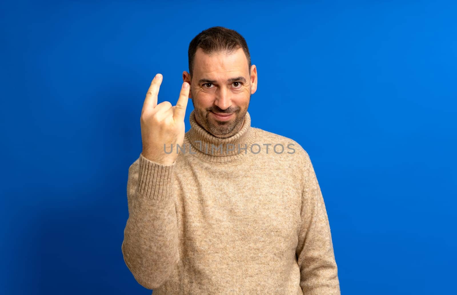 Portrait of bearded hispanic man showing rock and roll gesture holding heavy metal sign, enjoying favorite music at party, having fun exclaiming with joy. Indoor studio shot isolated blue background.