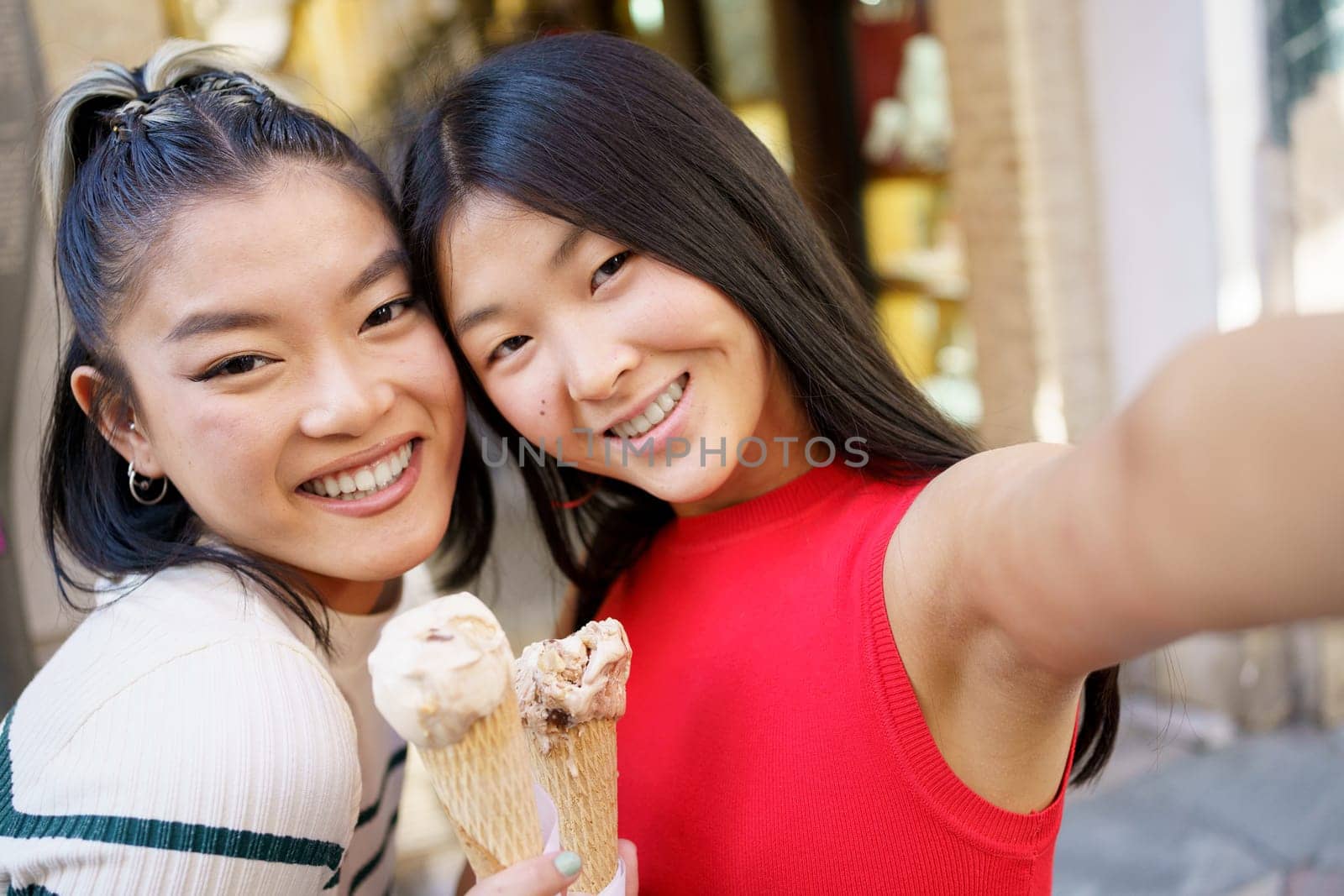 Positive young Asian women smiling happily and taking selfie while eating delicious ice cream cone together in street