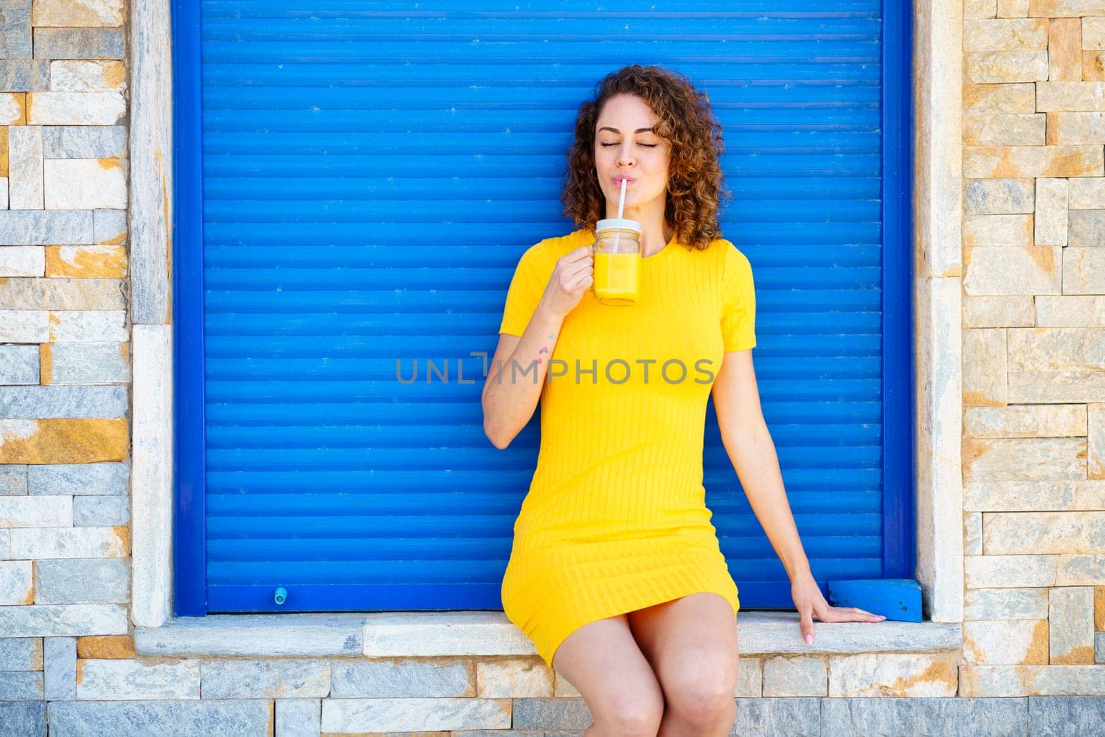 Charming young female in yellow dress with curly brown hair and closed eyes enjoying fresh cold juice while sitting on stone border on city street