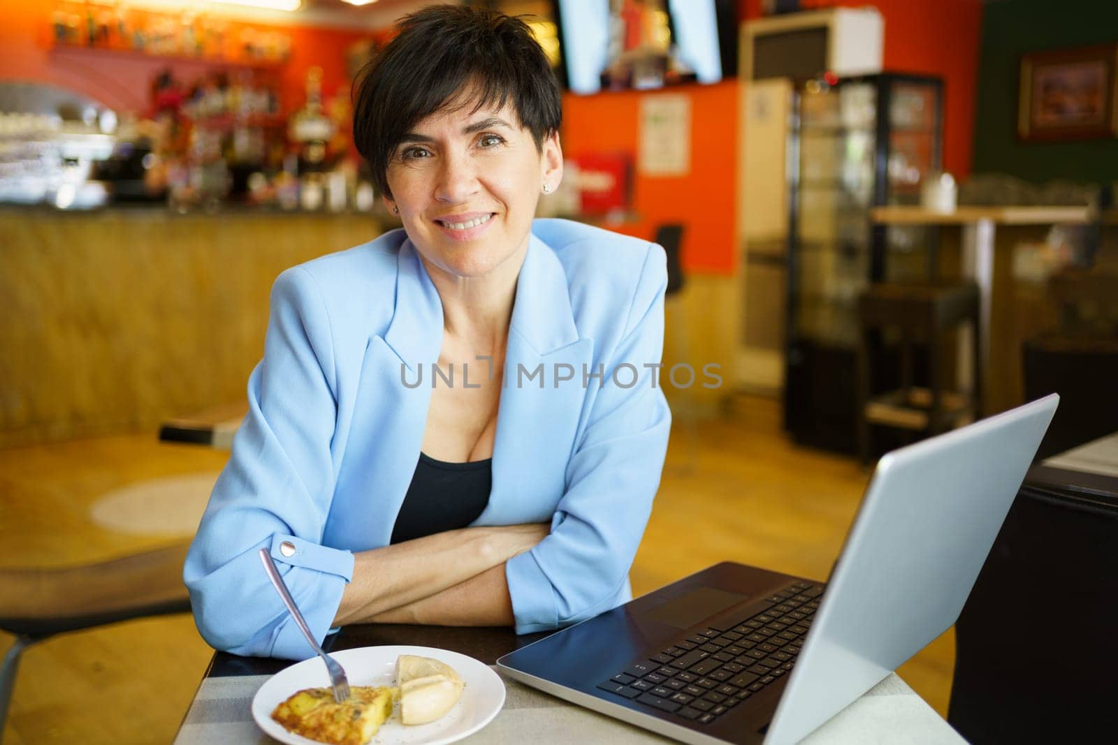 Smiling mature woman with food working on laptop in cafe by javiindy