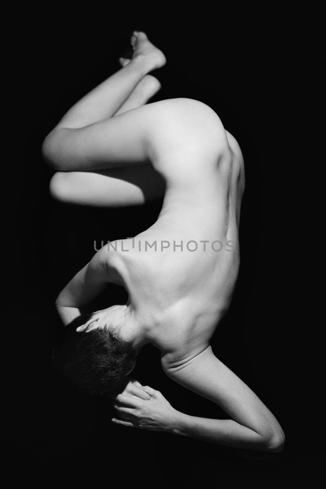 Full body of anonymous fit naked woman with short dark hair lying on floor against black background in studio. Black and white photograph
