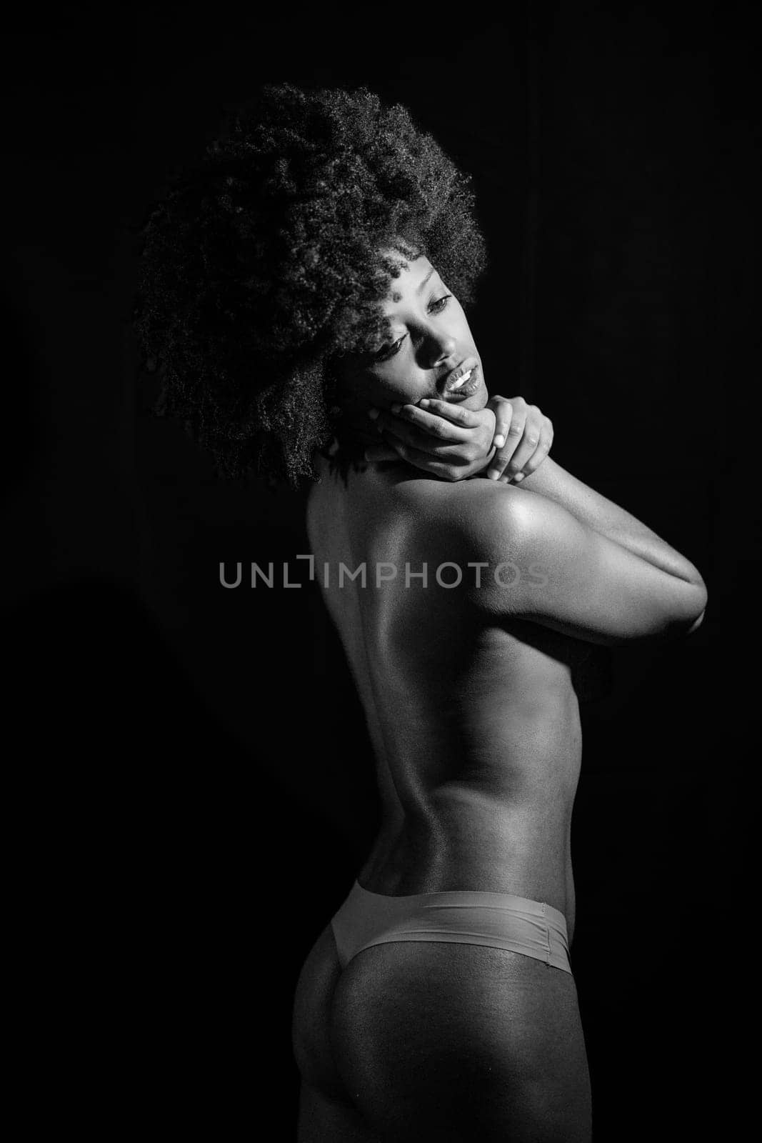 Sensual nude African American female with curly hair looking away over shoulder while standing against black background. Black and white photograph.