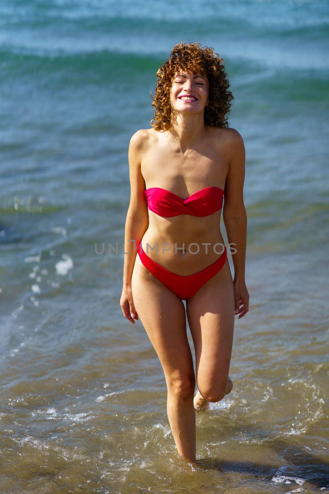 Smiling young female in pink swimwear with curly hair walking in ocean water while enjoying summer vacation on sunny day