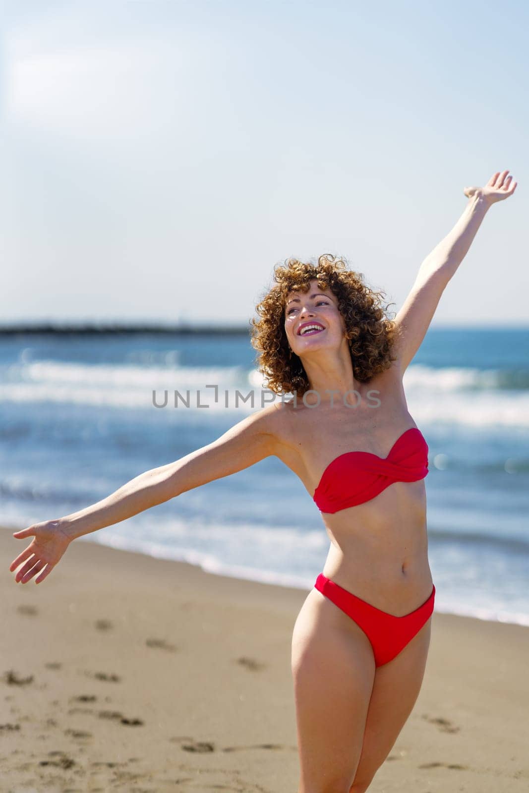 Cheerful young female with curly ginger hair in bikini, standing on sandy beach and smiling with stretched arms towards sky and earth in sunlight while looking away against blurred seawater