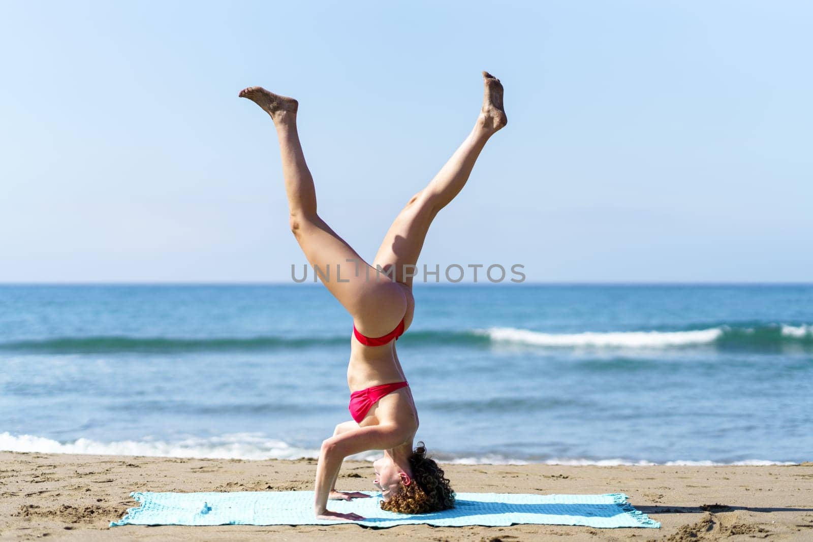 Side view of young slim lady in bikini doing Tripod Headstand with legs apart on sandy coastline of ocean