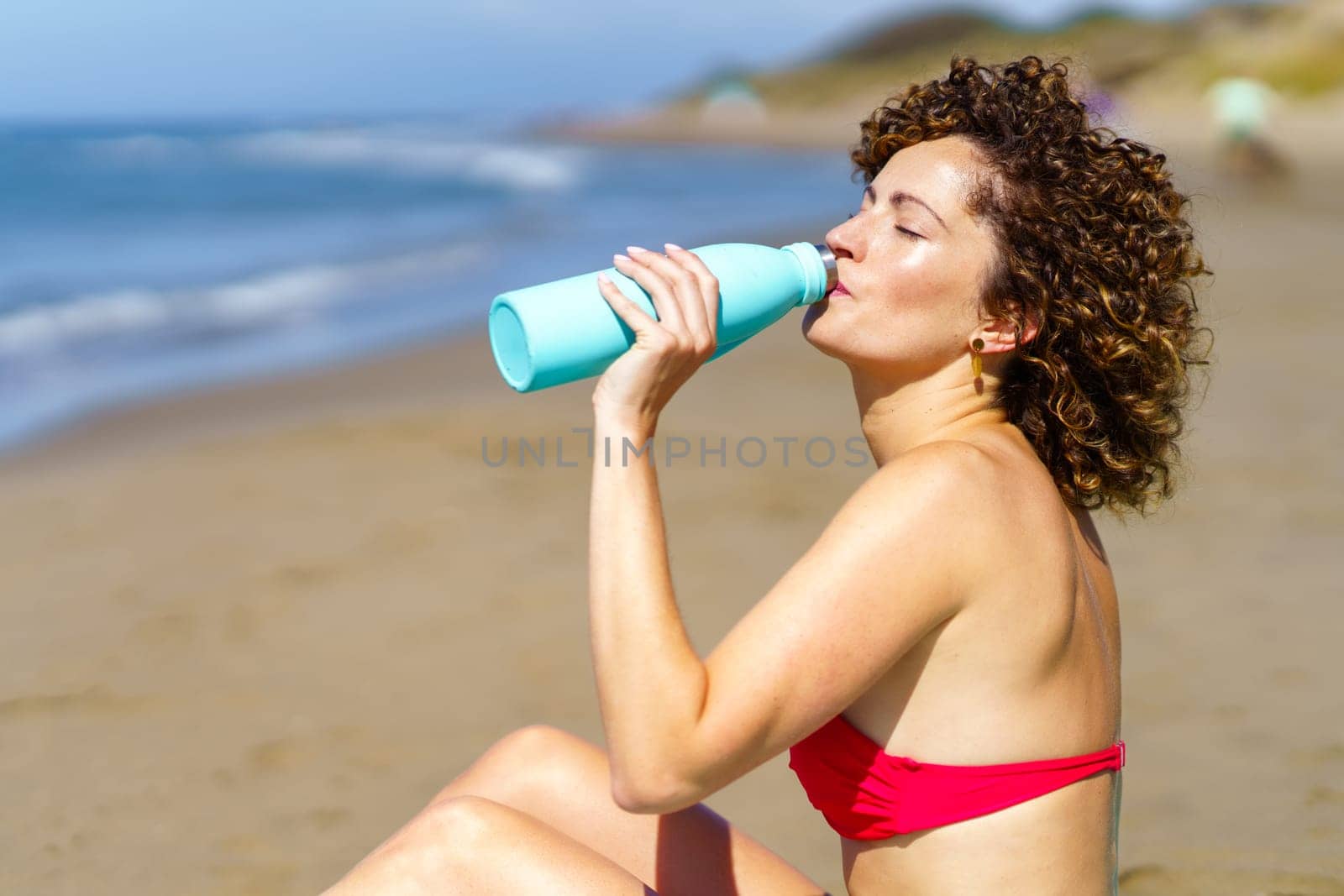 Side view of young redhead female in bikini bra drinking refreshing water from bottle while sunbathing on coast