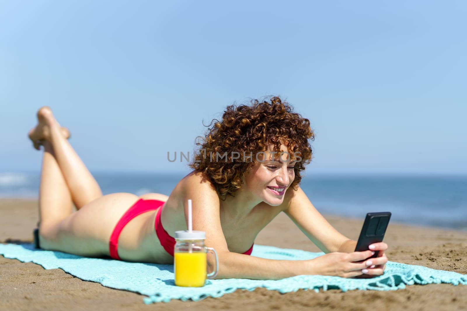 Happy woman with ginger curly hair in red bikini lying on coast of ocean with refreshing drink and texting on smartphone