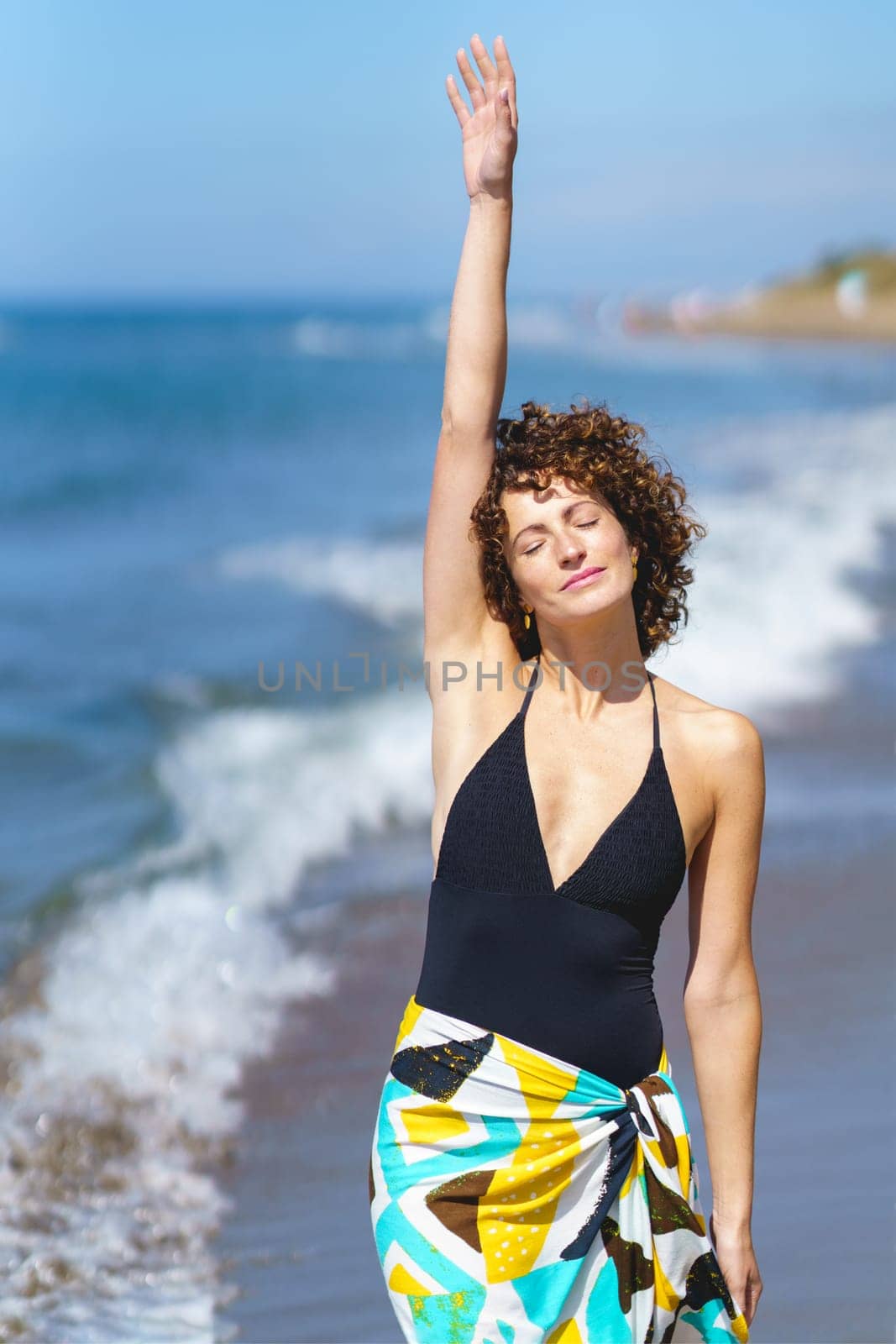 Positive young female in black swimsuit and colorful casual cloth, standing with raised arm and closed eyes on blurred background of sea and blue sky in daylight