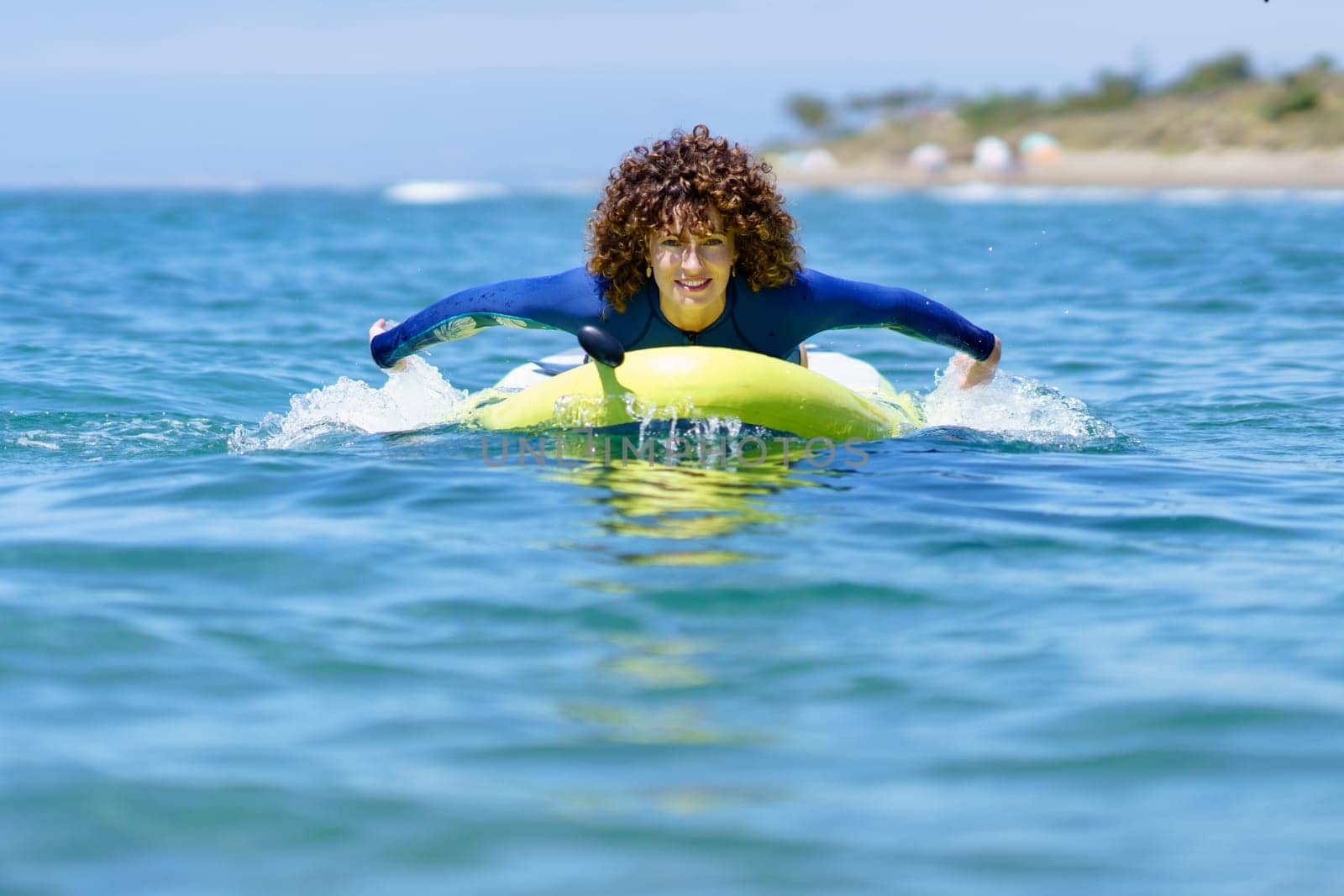 Content curly haired female in wetsuit pulling SUP board in rippling seawater and looking at camera in daylight