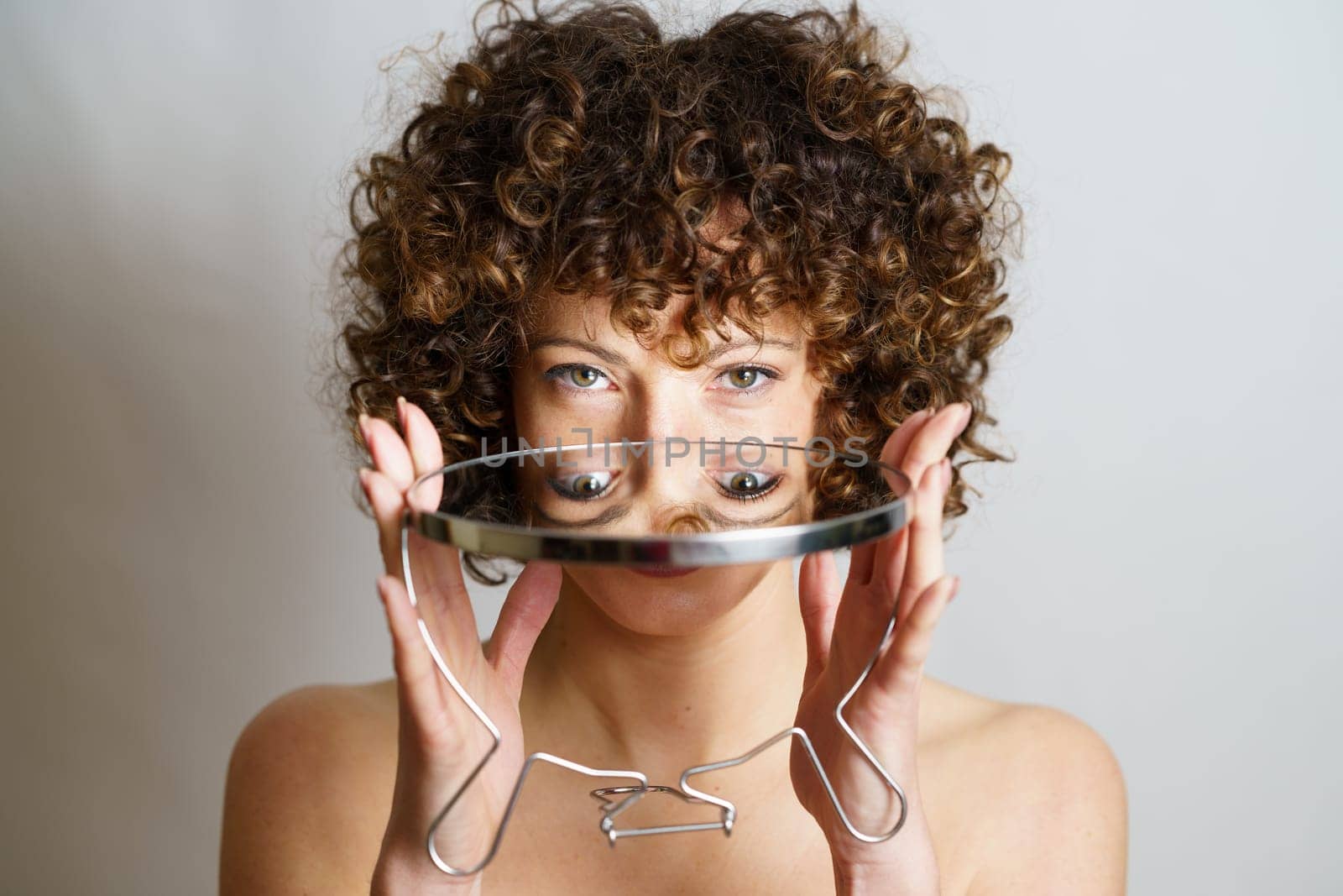 Attractive young curly haired girl model holding round mirror in hands with reflection of face and eyes while looking at camera against gray background