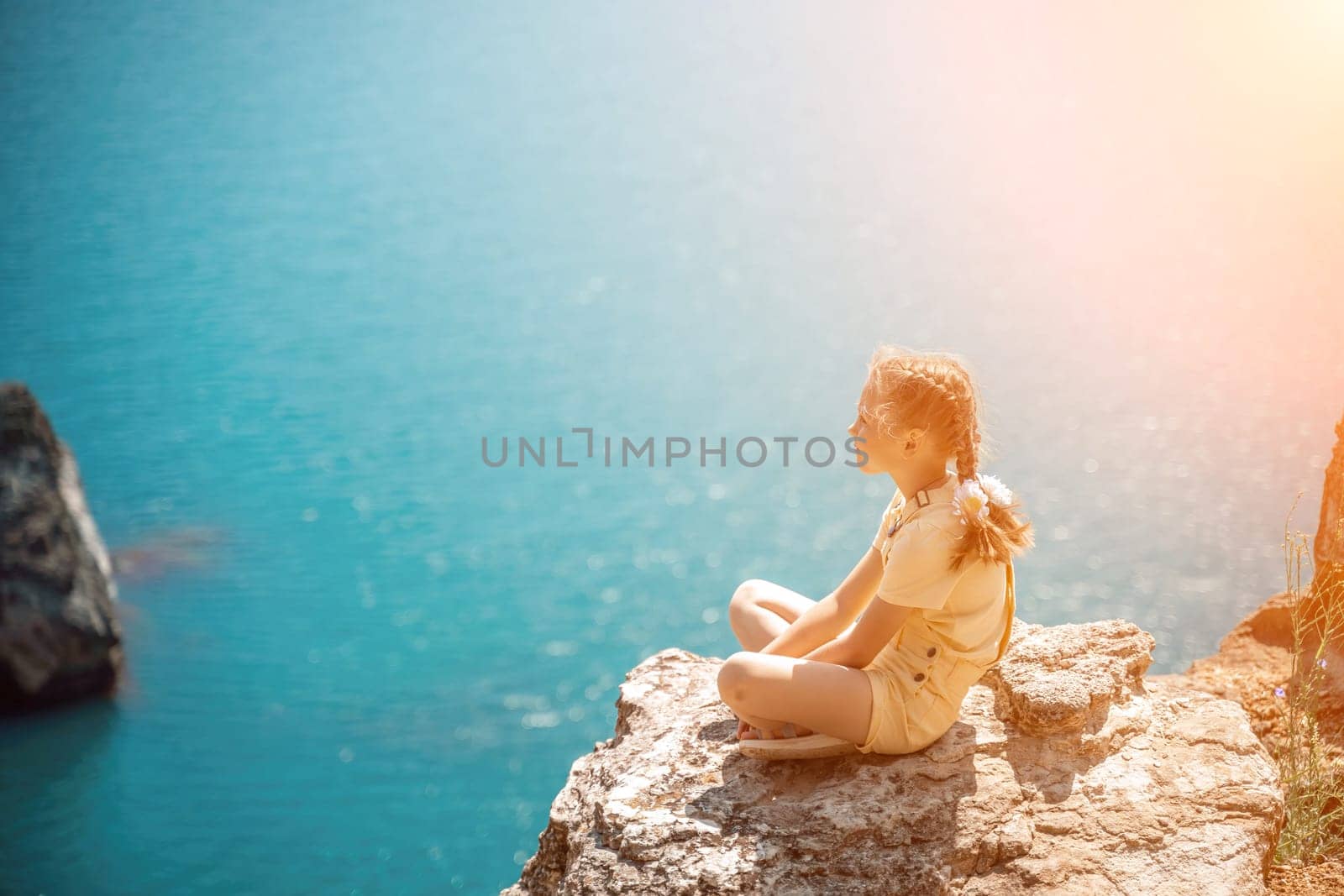 Happy girl perched atop a high rock above the sea, wearing a yellow jumpsuit and braided hair, signifying the concept of summer vacation at the beach. by Matiunina