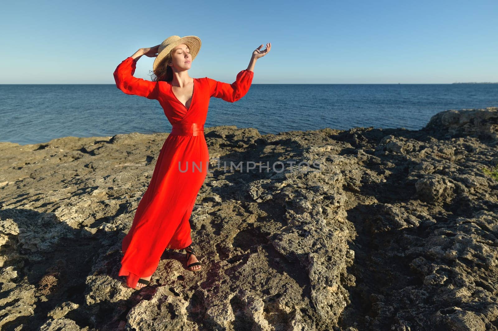 Young beautiful woman in red dress standing looking at the sea. Girl in a straw hat in nature against the blue sky and the ocean. Fashion photo, renaissance-style pose.