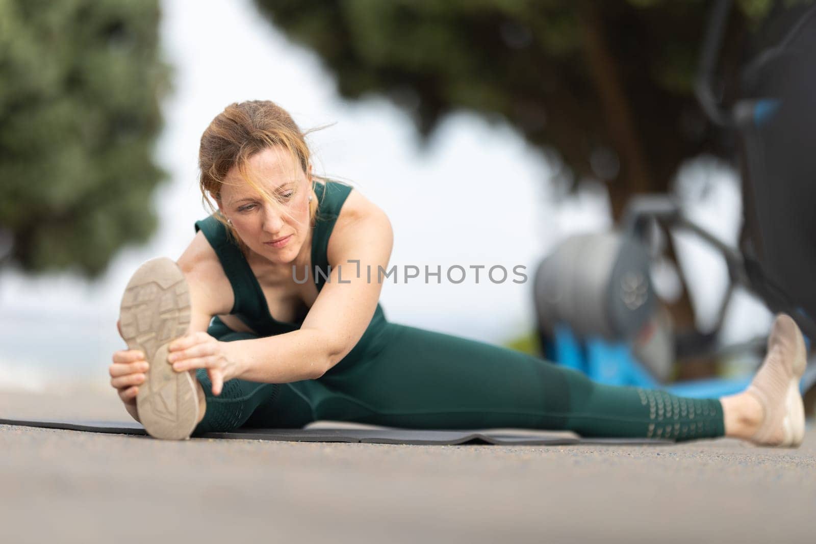 Adult sportive woman sitting in a splits at the outdoor sports ground and stretching to the side to her leg. Mid shot