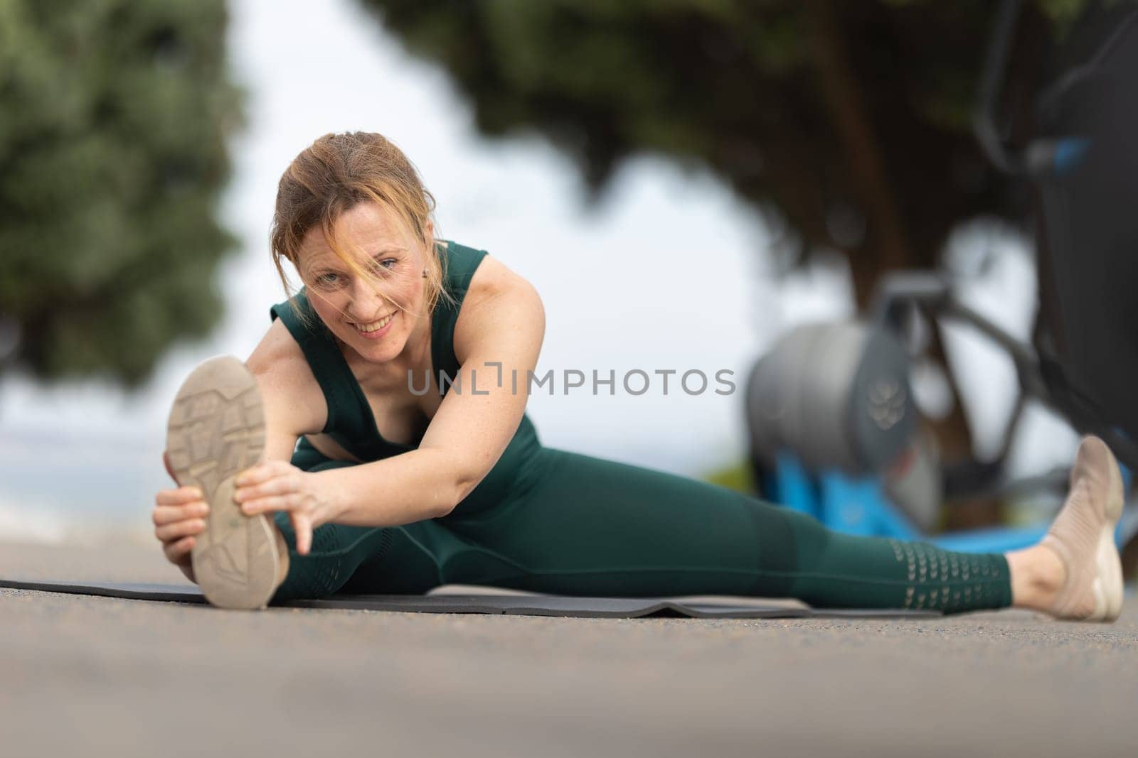 Adult smiling woman sitting in a splits at the outdoor sports ground and stretching down to her leg. Mid shot