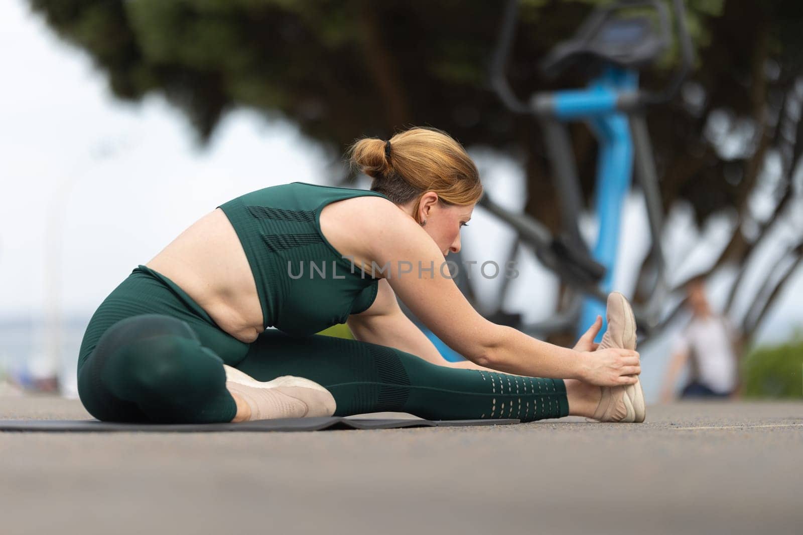 Adult woman sitting in a splits at the outdoor sports ground and stretching down to her leg. Mid shot
