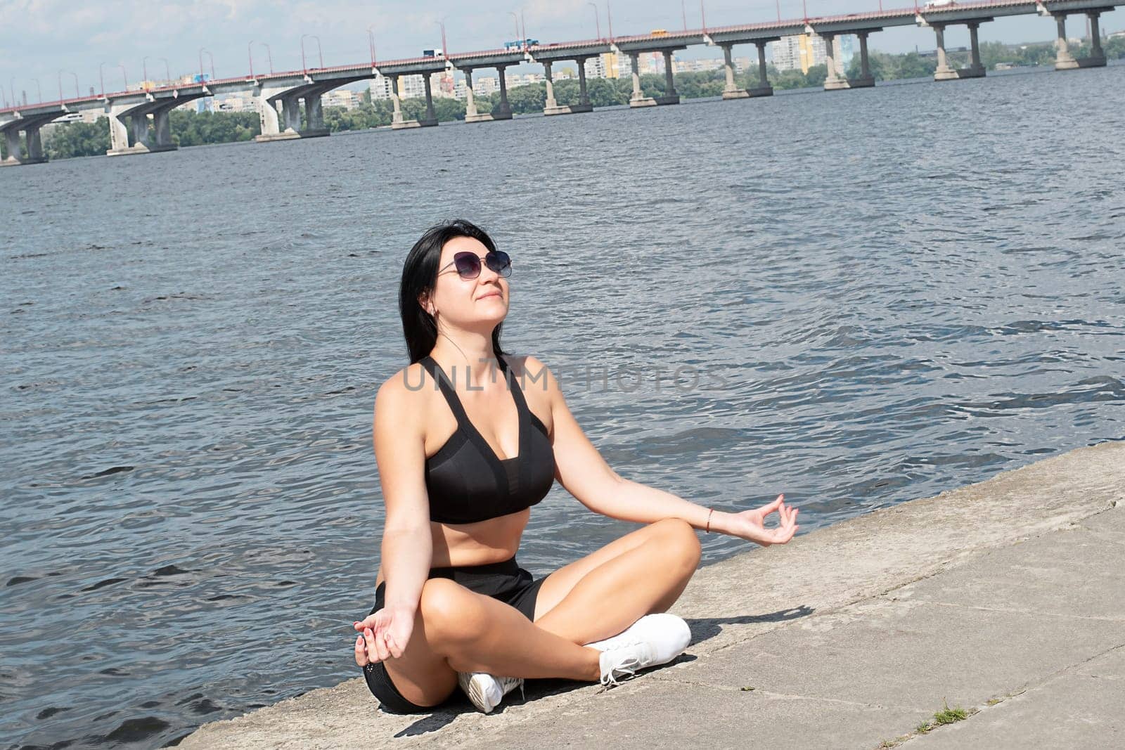 Yoga concept. A beautiful girl in a lotus position sits on the embankment near the river in the street in the fresh air. Meditate with closed eyes.