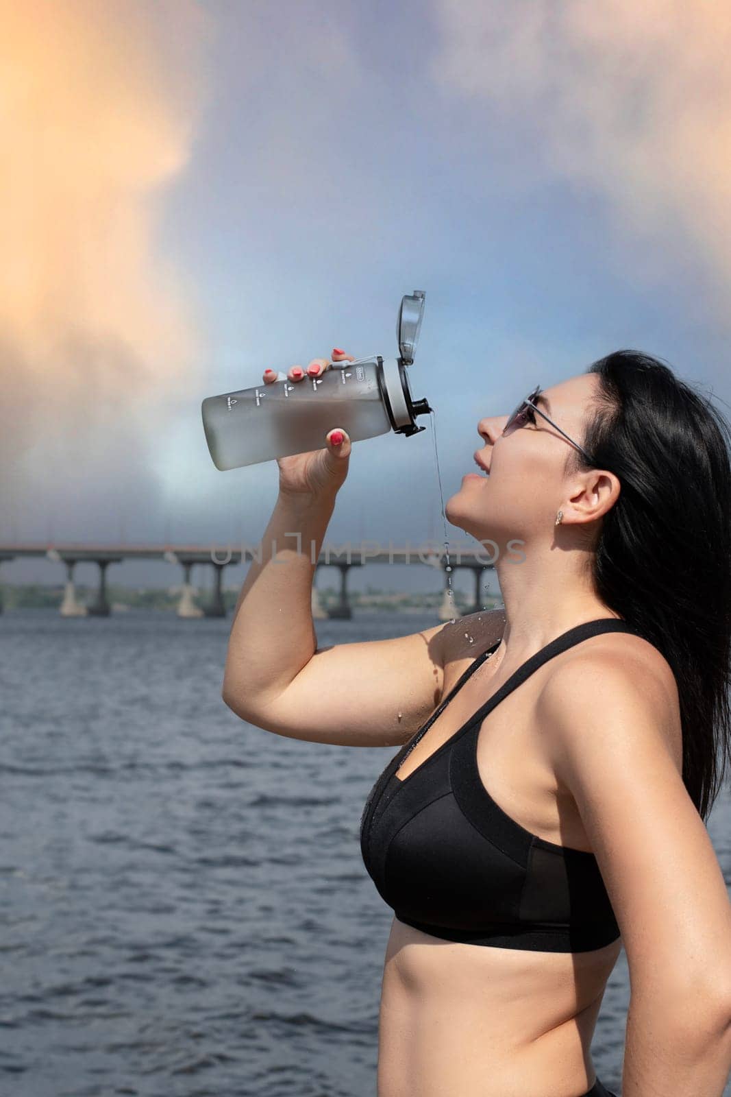 A beautiful and slender girl in sunglasses pours water over herself from a sports bottle after a fitness workout on the street in the summer, against the sky with a rainbow. Sport concept. Close-up.