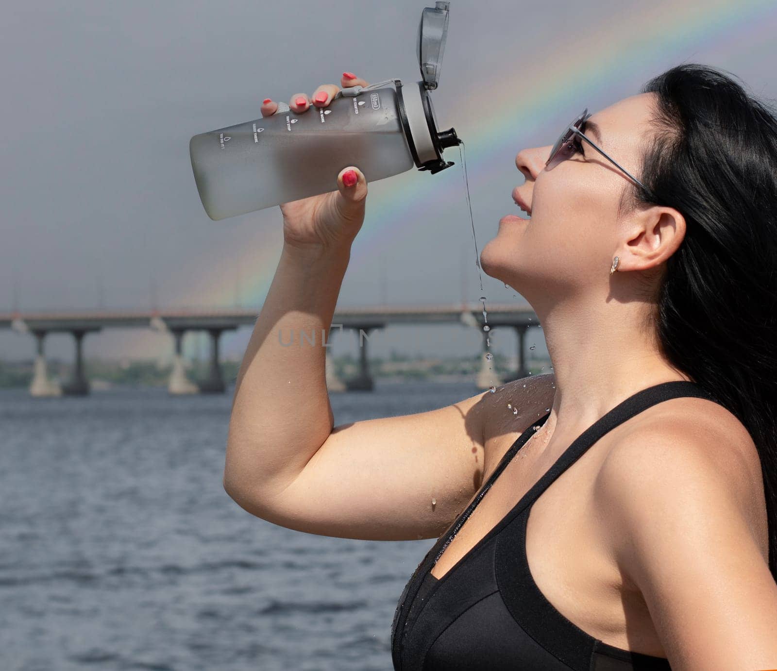 A beautiful and slender girl in sunglasses pours water over herself from a sports bottle after a fitness workout on the street in the summer, against the sky with a rainbow. Sport concept. Close-up.
