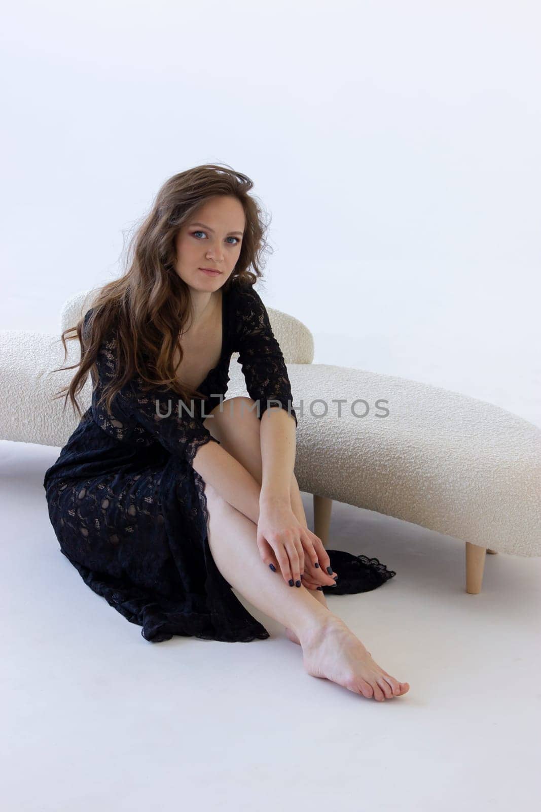 Young Attractive Girl Sits Near Beige Sofa On White Background. Beautiful Caucasian Woman With Long Brown Hair Looks At Camera. Modern Furniture. Vertical Plane. High quality photo