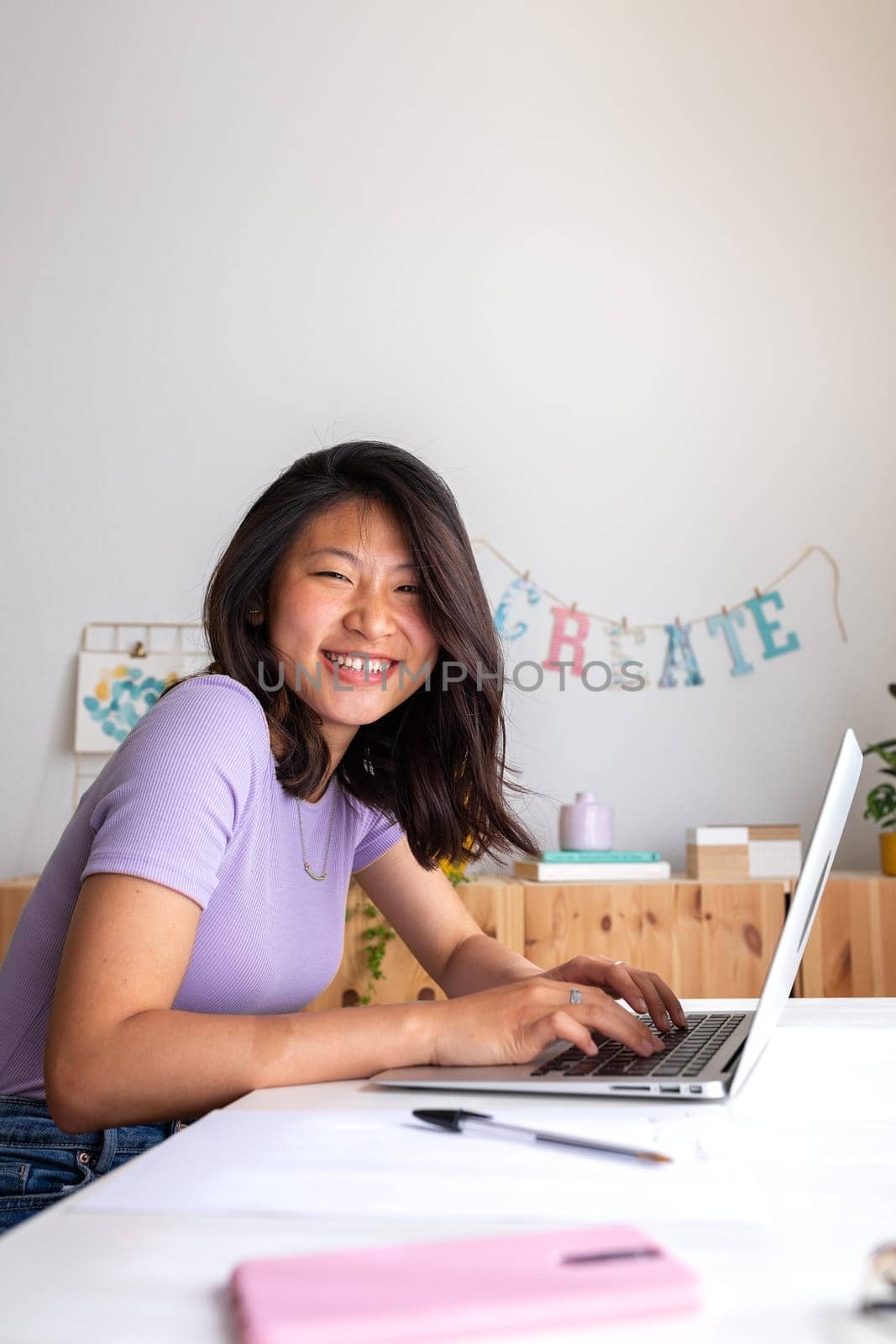 Vertical portrait of happy, smiling teen asian female college student studying at home, doing homework using laptop looking at camera. Copy space. Education, e-learning concept.