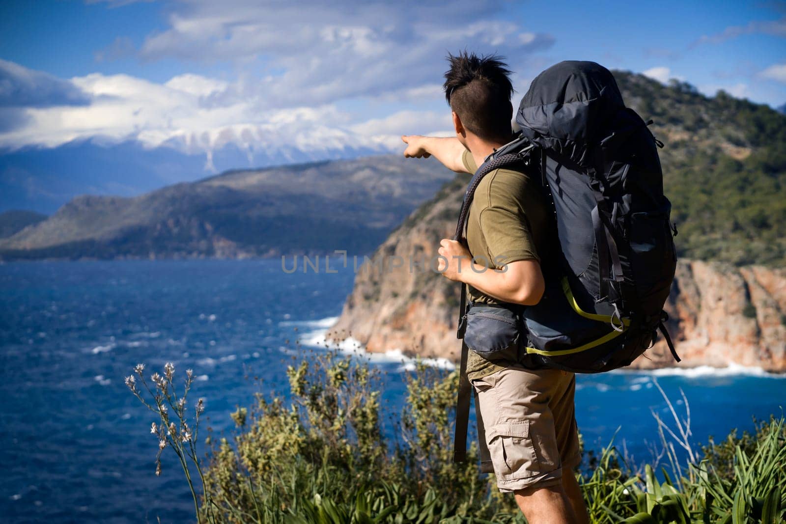 A young man is traveling, trekking and walking along the sea coast on a sunny day with a backpack. The traveler points with his hand to a beautiful mountain on the horizon.