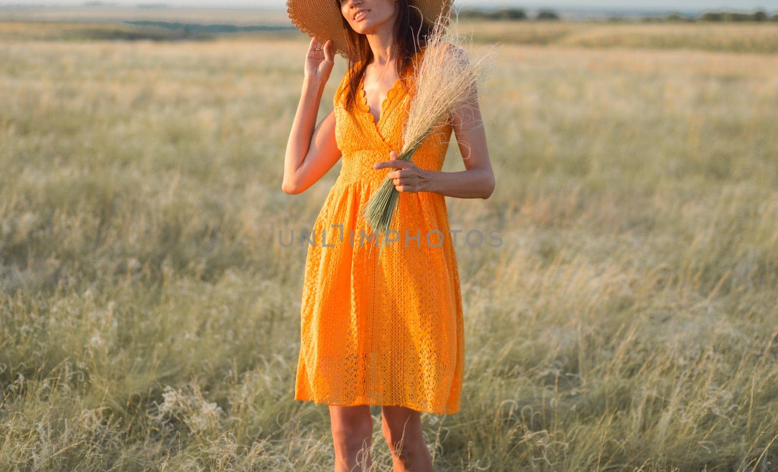 Young woman in an orange dress and a straw hat standing on a field in the rays of the setting sun by Ekaterina34