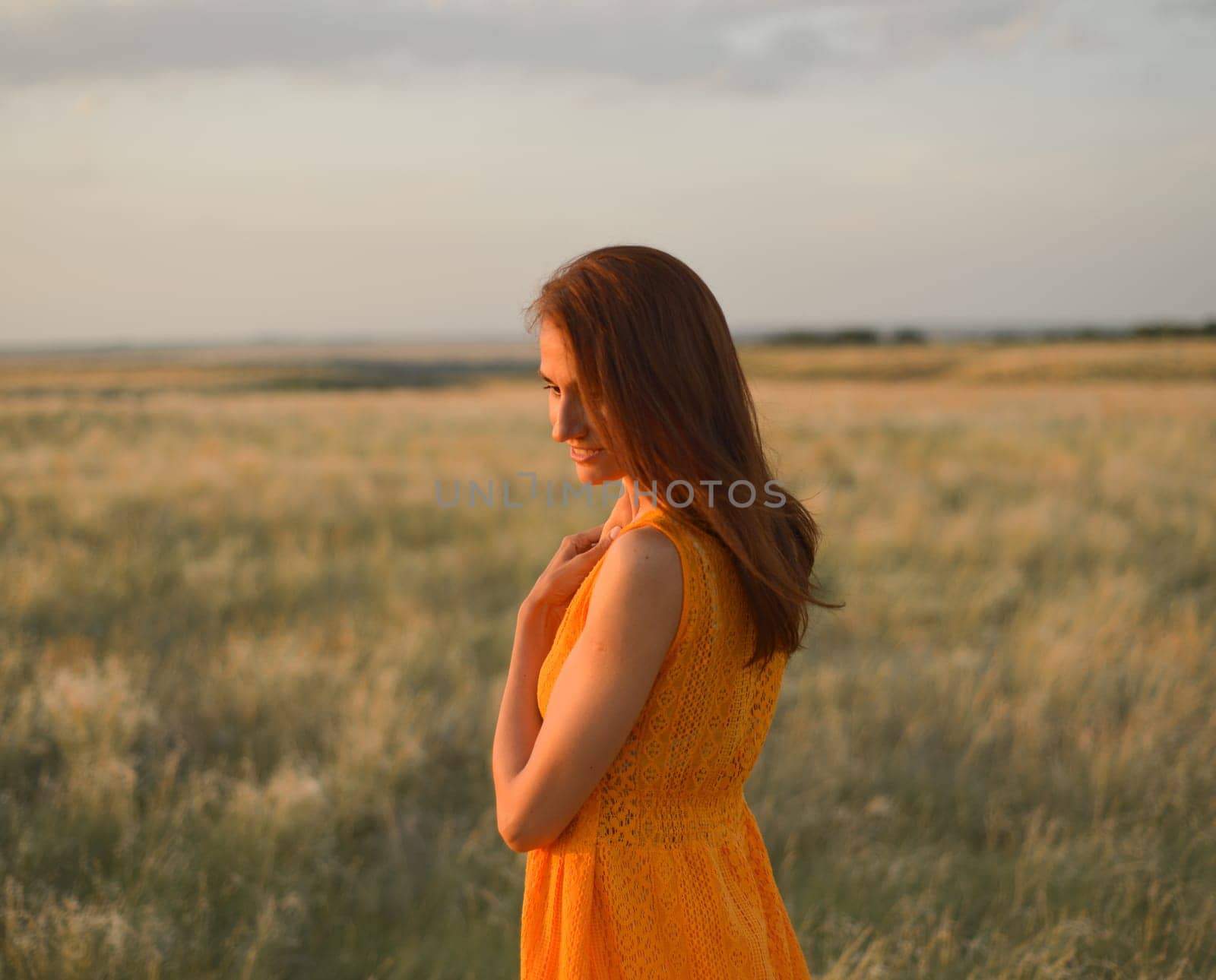 A pretty young girl with long brown hair in an orange dress in a field at sunset. The concept of freedom and nature. by Ekaterina34
