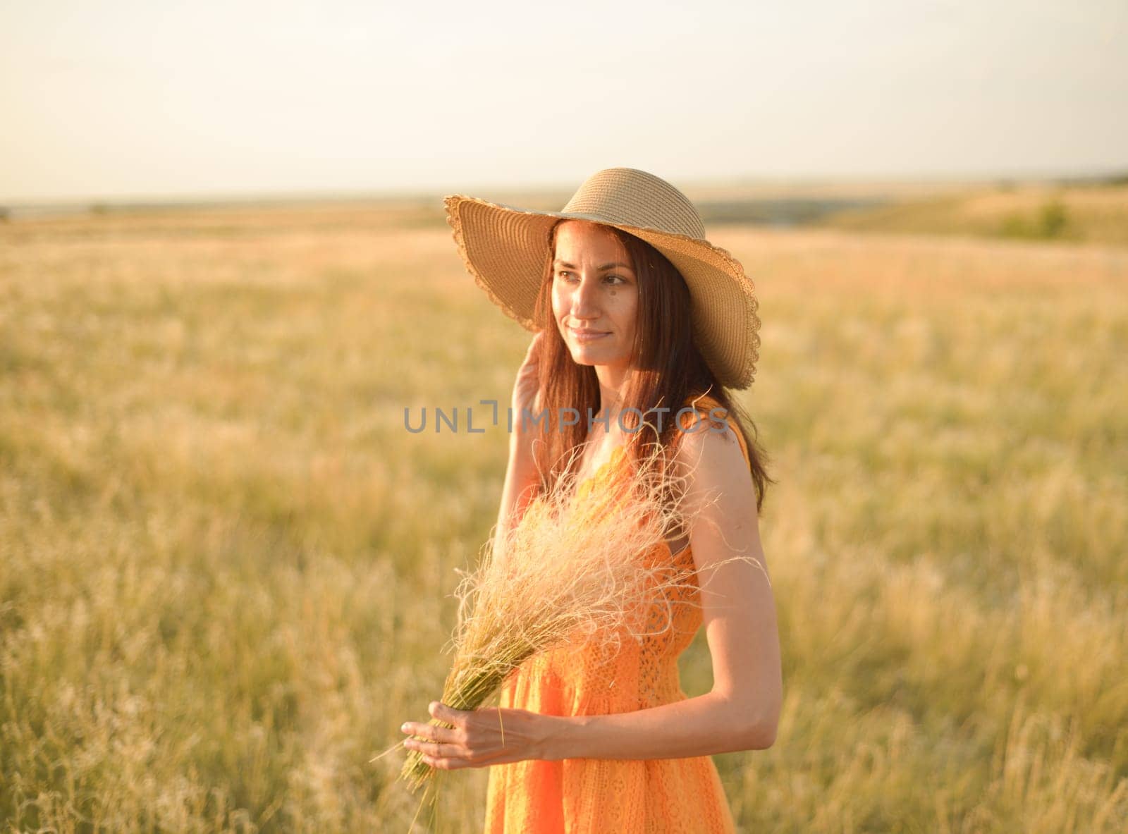 Young woman in an orange dress and a straw hat standing on a field in the rays of the setting sun by Ekaterina34