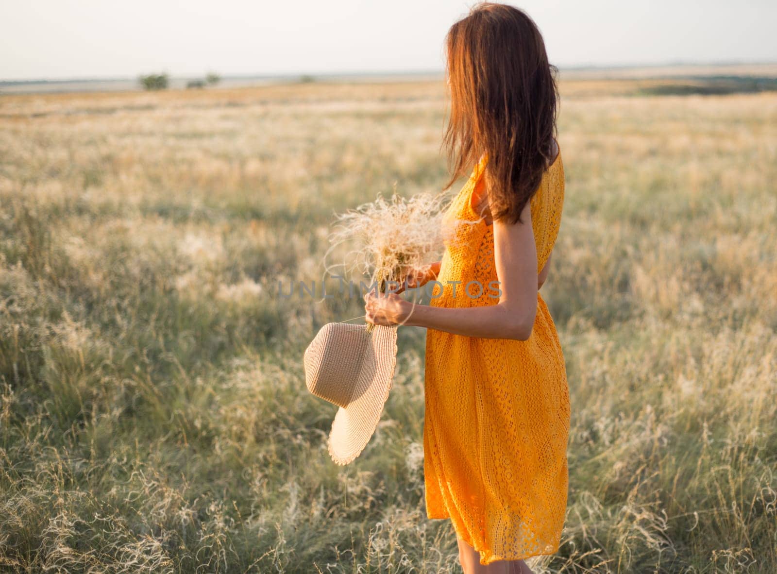Beauty romantic girl outdoors. Rear view of a beautiful girl dressed in a casual orange dress with a straw hat in her hands on a field in the sunlight. Blows long hair. Autumn. Shine the sun, sunshine. backlit Warm tinted