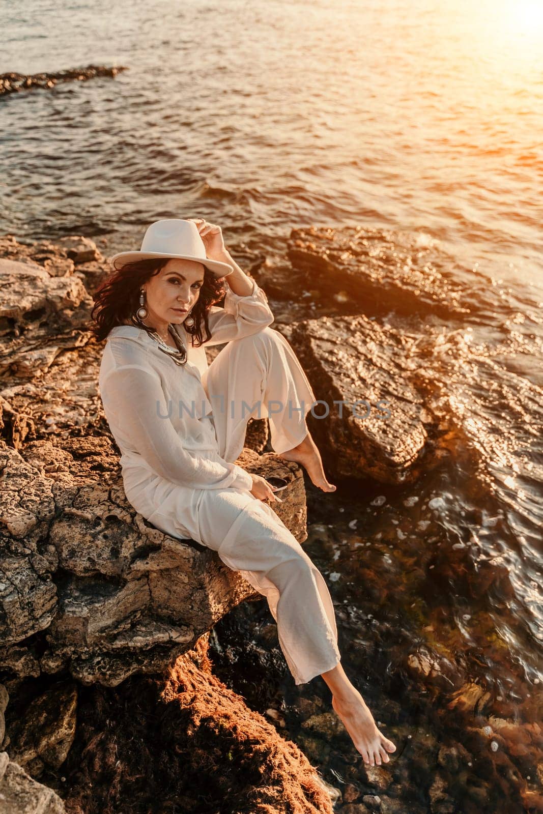 woman sea sunset. woman in a white pantsuit and hat is sitiing on the beach enjoying the sea. Happy summer holiday.