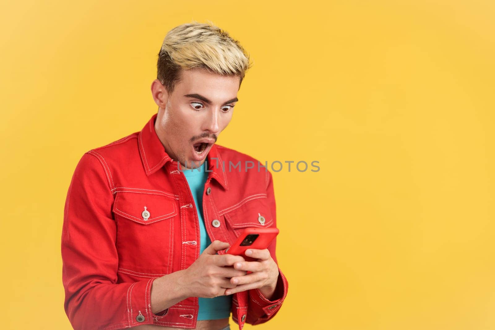 Surprised gay man looking the screen of a mobile phone in studio with yellow background