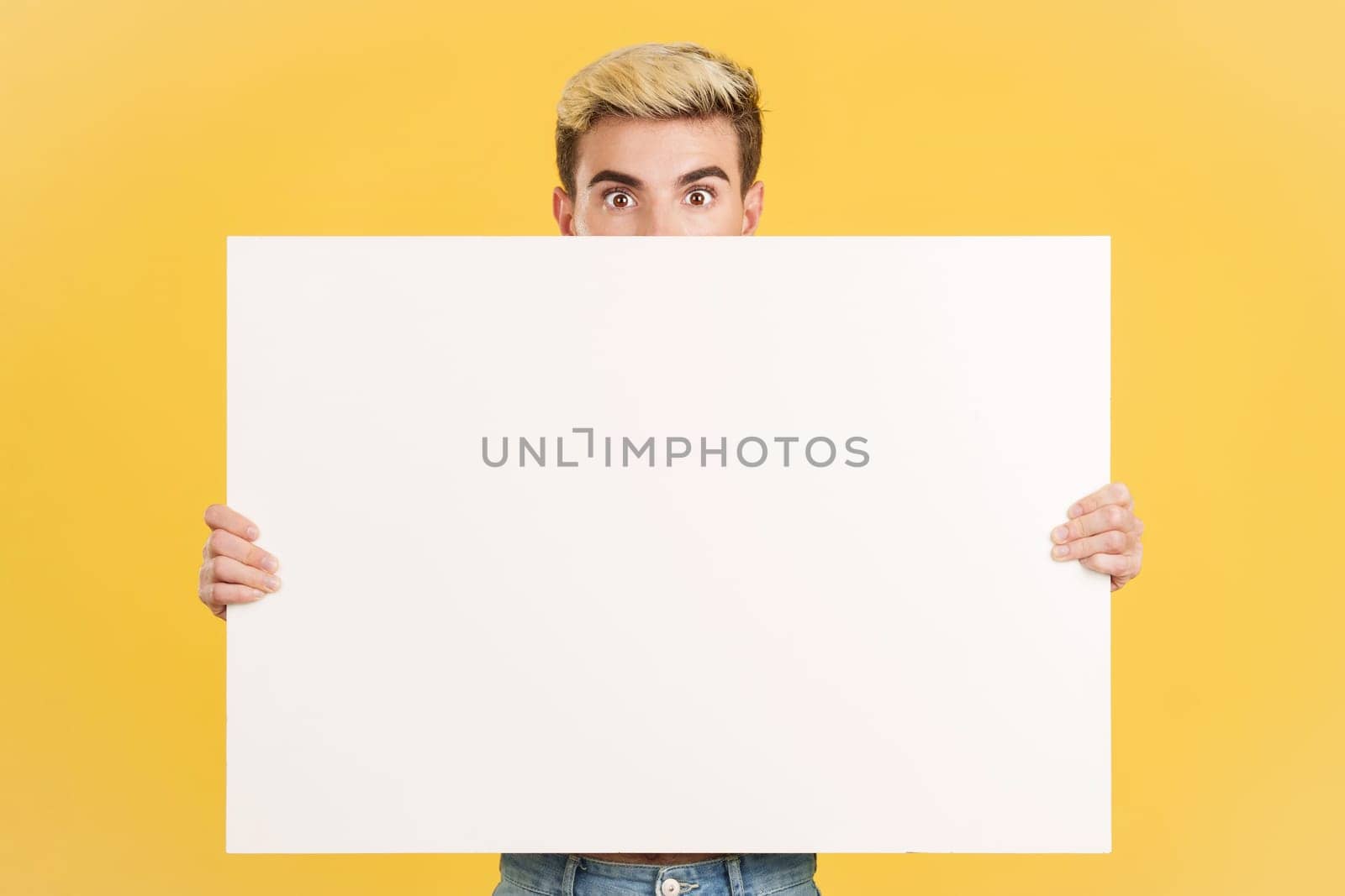 Surprised gay man hiding behind a blank board in studio with yellow background