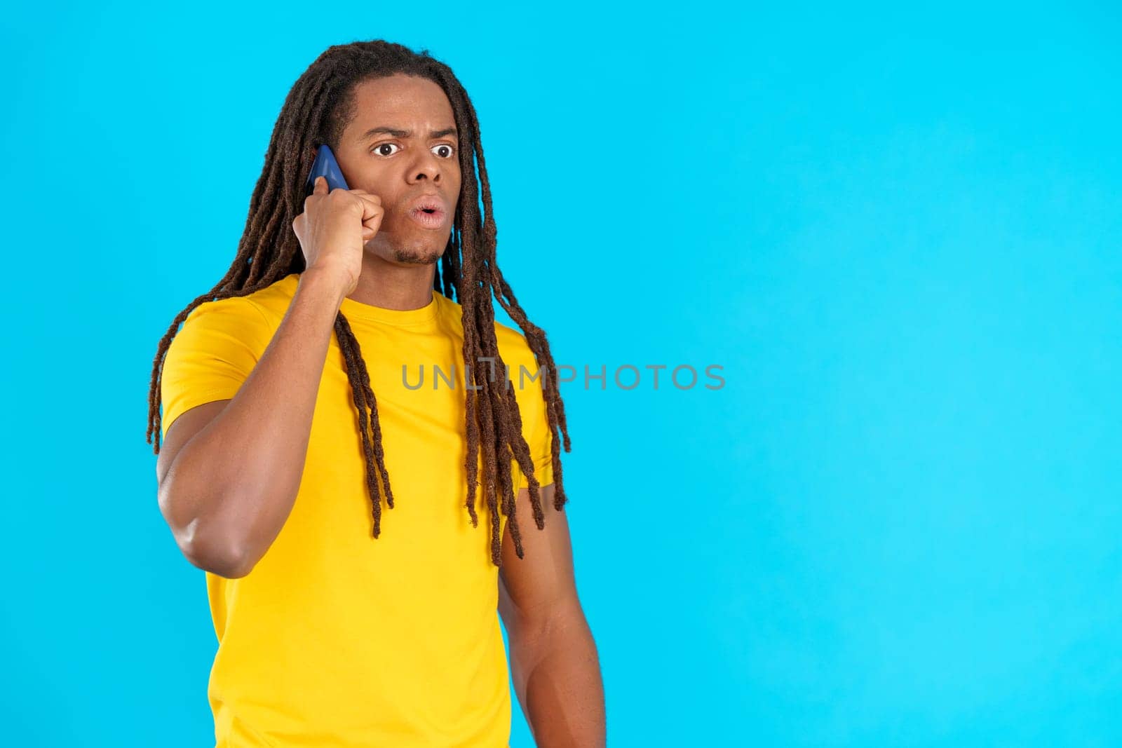 Surprised latin man with dreadlocks talking to the mobile in studio with blue background