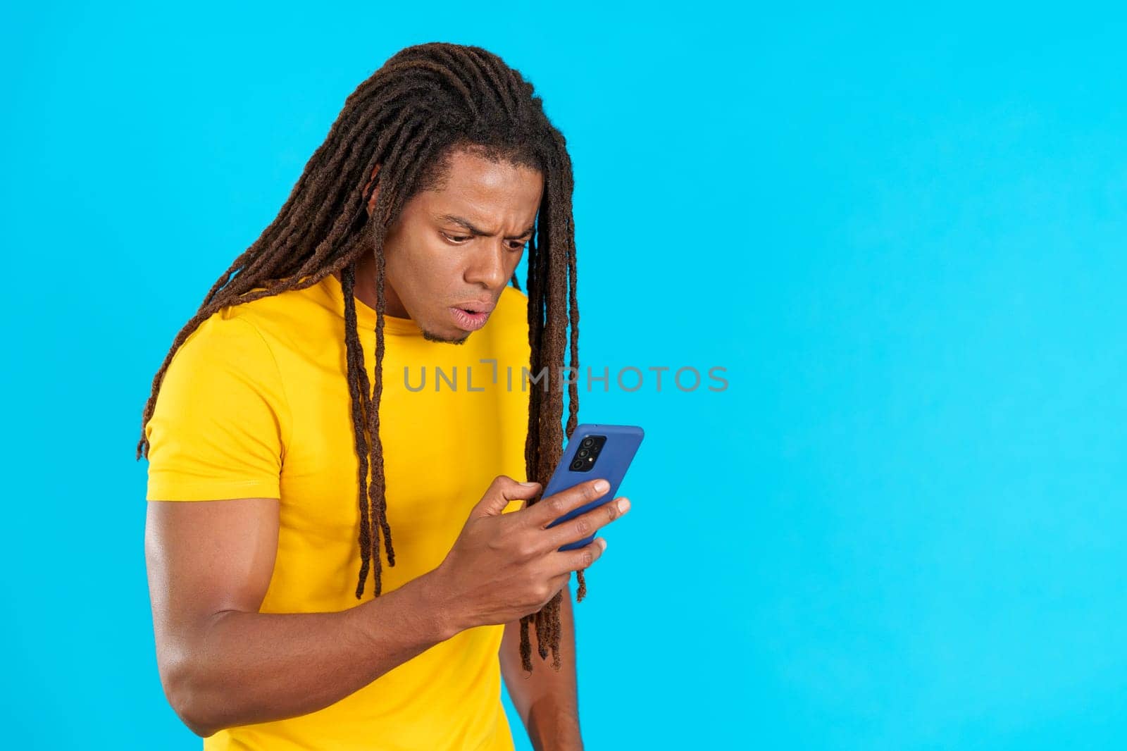 Surprised latin man with dreadlocks looking the screen of a mobile in studio with blue background