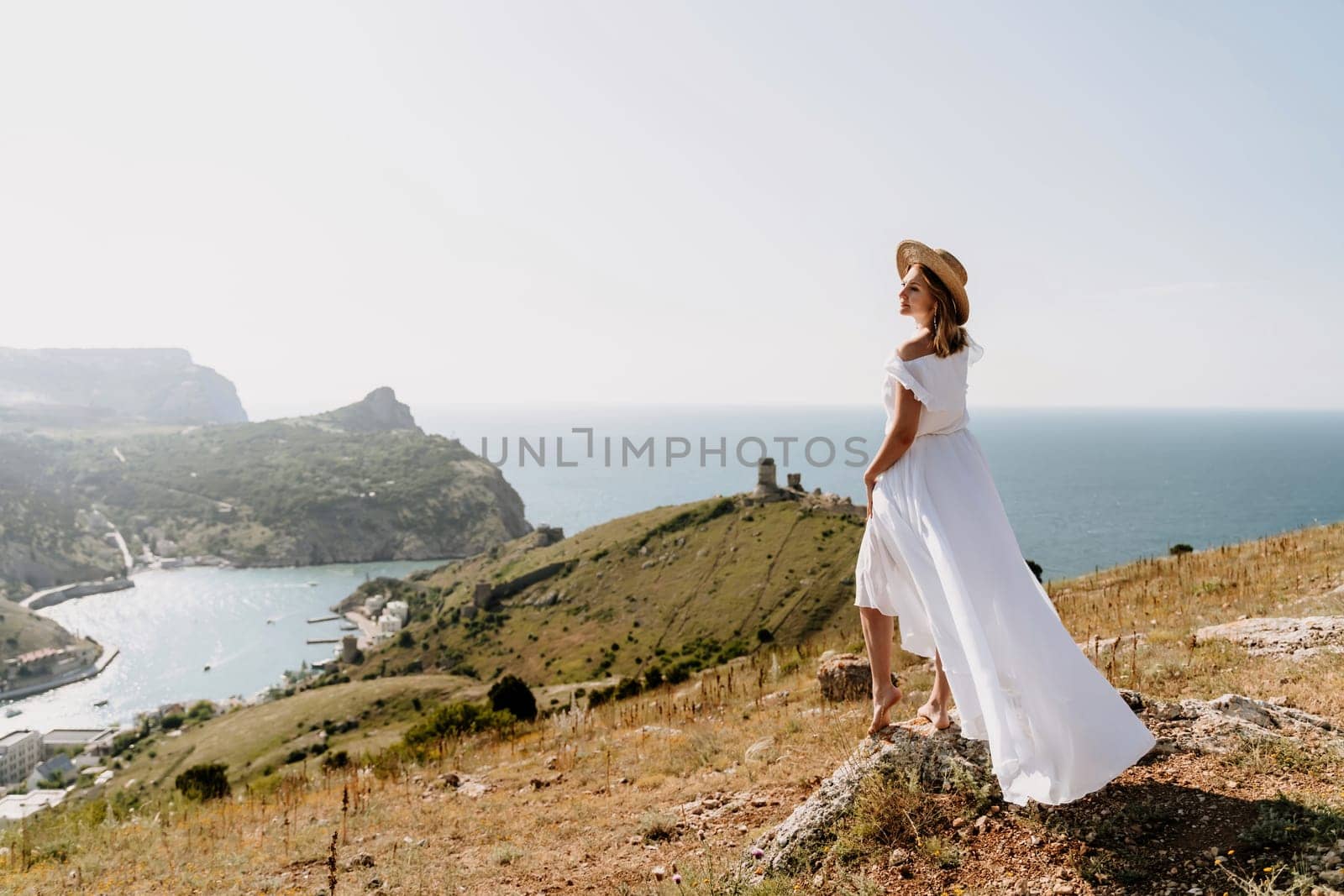 Happy woman in a white dress and hat stands on a rocky cliff above the sea, with the beautiful silhouette of hills in thick fog in the background