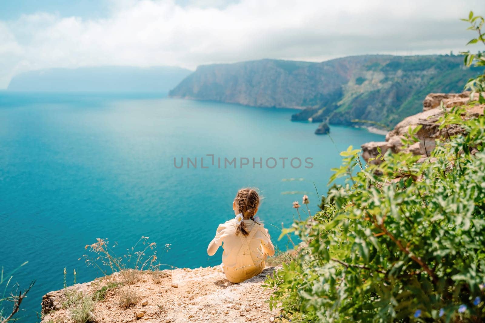 Happy girl perched atop a high rock above the sea, wearing a yellow jumpsuit and braided hair, signifying the concept of summer vacation at the beach