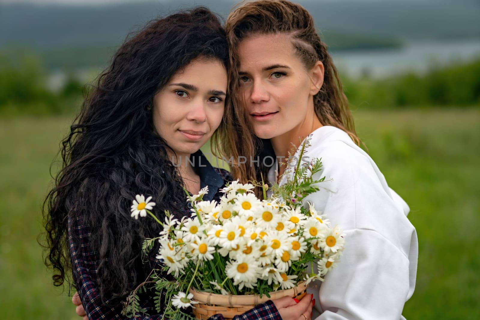 Two women enjoy nature in a field of daisies. Girlfriends hugging hold a bouquet of daisies and look at the camera. by Matiunina