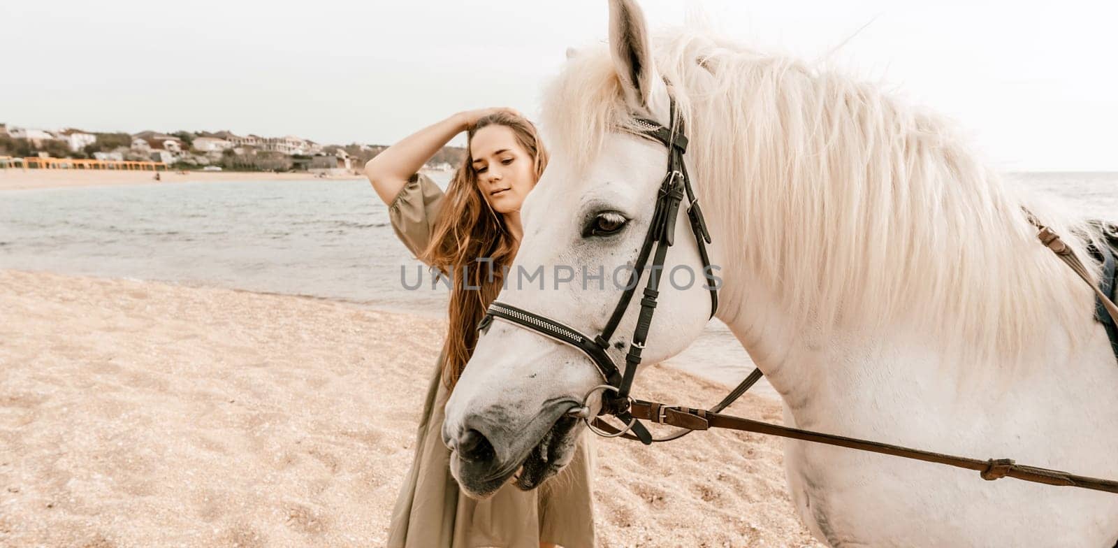 A woman in a dress stands next to a white horse on a beach, with the blue sky and sea in the background