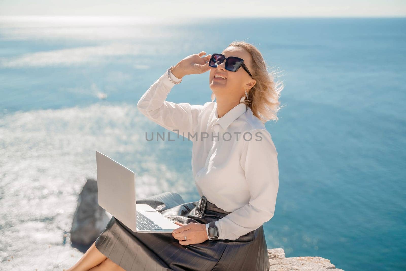 Business woman on nature in white shirt and black skirt. She works with an iPad in the open air with a beautiful view of the sea. The concept of remote work