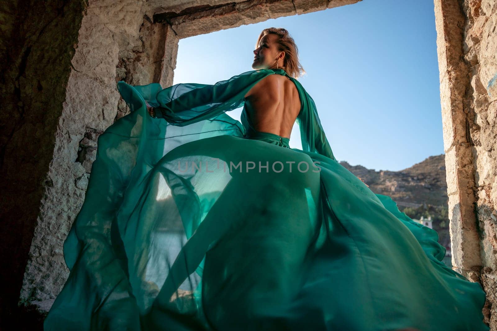 Rear view of a happy blonde woman in a long mint dress posing against the backdrop of the sea in an old building with columns. Girl in nature against the blue sky. by Matiunina