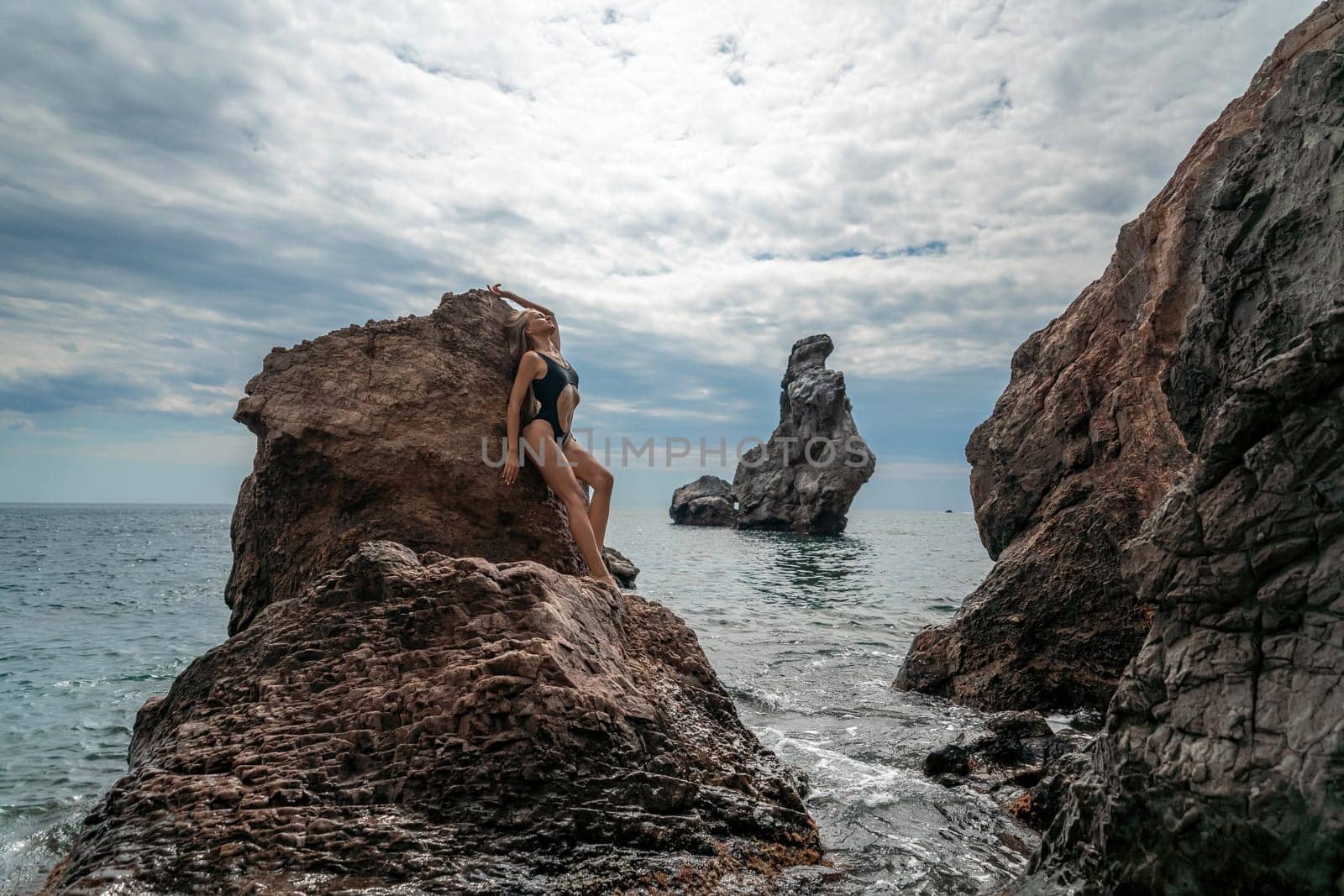 Woman swimsuit sea. Attractive blonde woman in a black swimsuit enjoying the sea air on the seashore around the rocks. Travel and vacation concept. by Matiunina