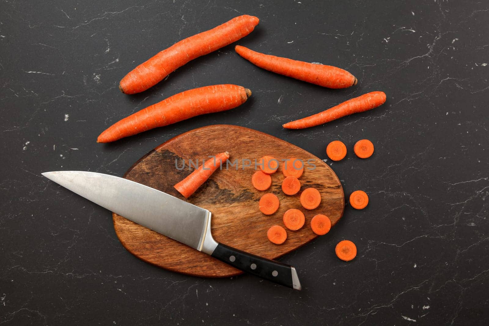 Top down view, chopping board, chef knife, and carrots, some of them cut in small circles, on black marble desk.