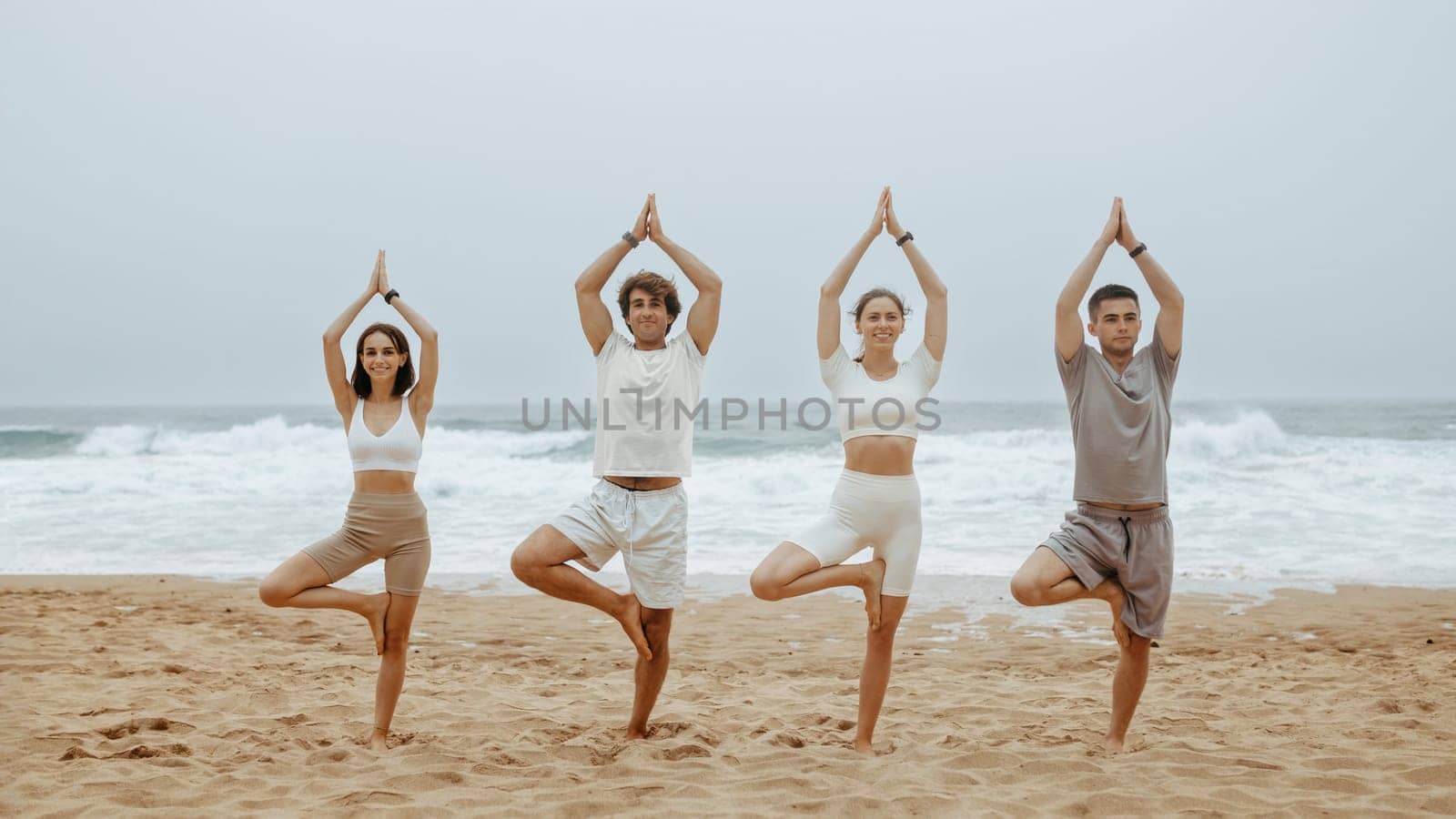 Healthy lifestyle concept. Group of men and women practising yoga, standing in tree pose on beach, having morning workout on ocean shore, full length