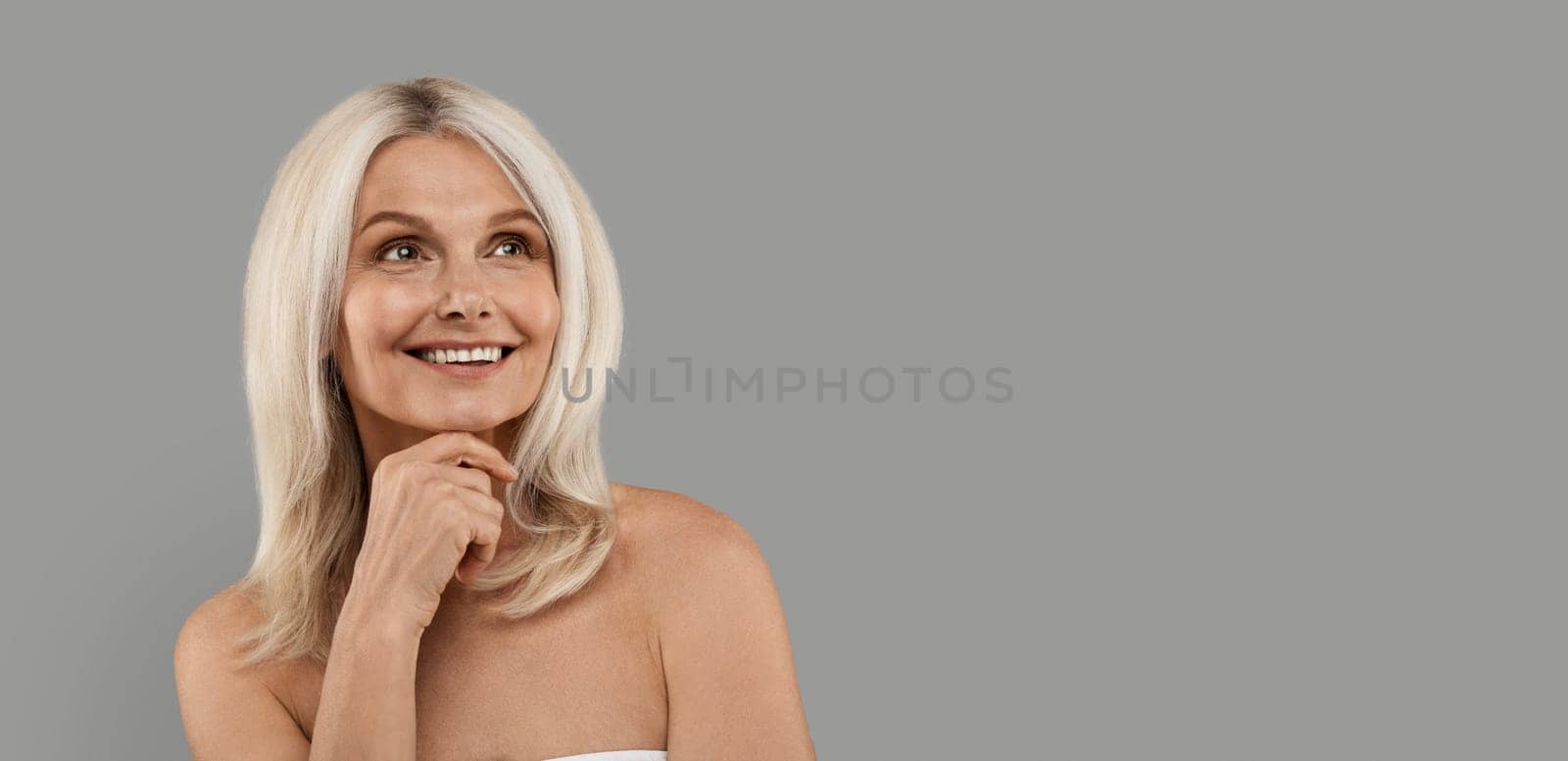 Beauty Shot Of Attractive Mature Woman Looking At Copy Space With Interest, Smiling Beautiful Senior Woman Wrapped In bath Towel Standing Over Grey Studio Background, Panorama With Free Place