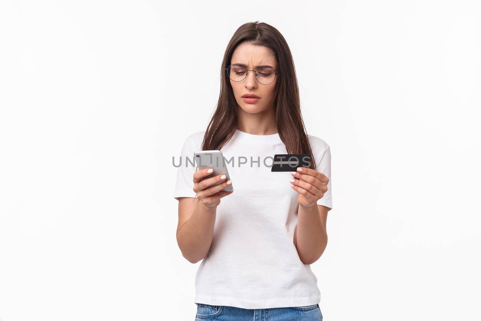 Portrait of serious-looking, focused young attractive woman copying digit numbers from credit card into billing info in app, paying or products online, purchase in internet store with smartphone.