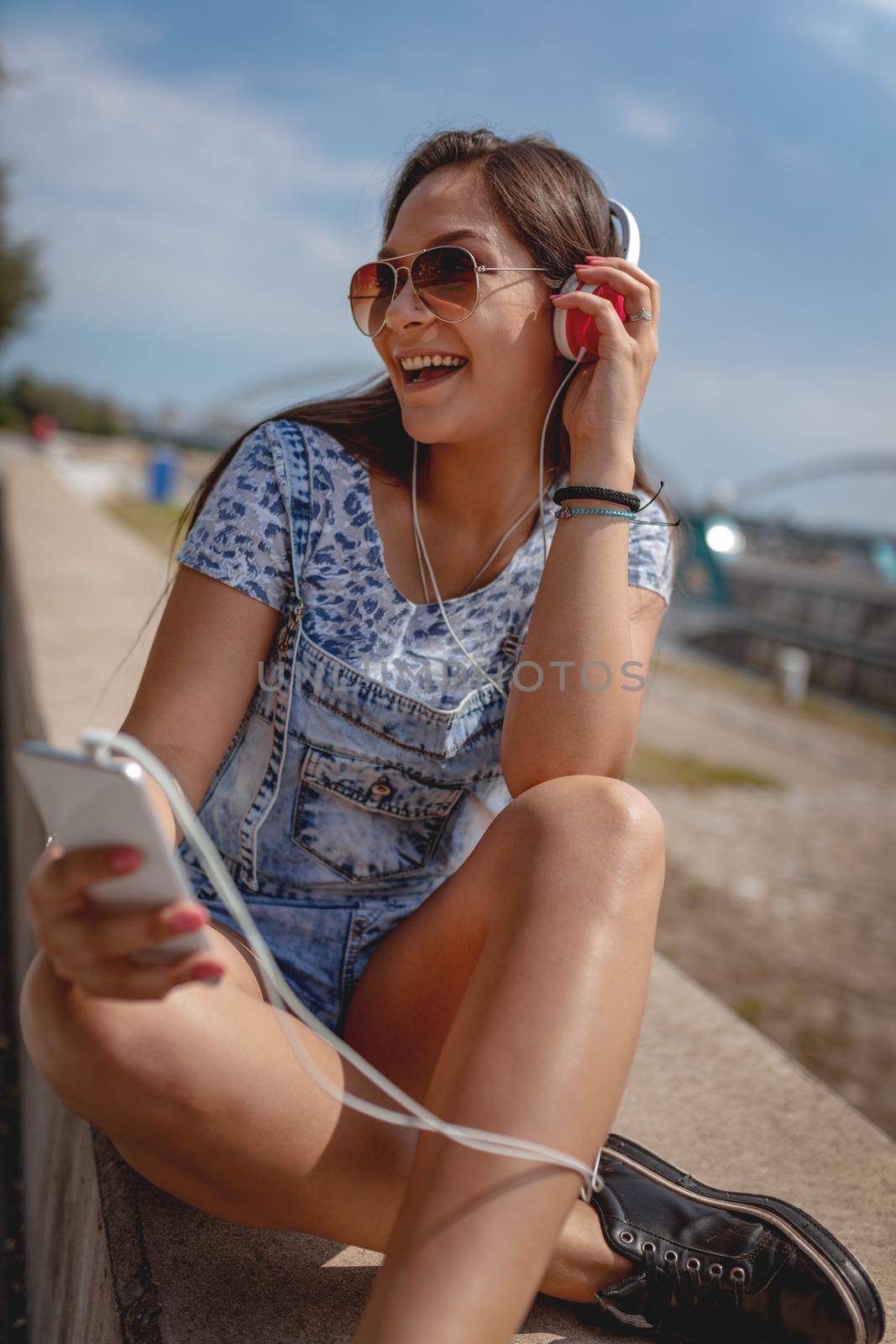 A beautiful cheerful young woman listening music from your phone and enjoying on the city rivershore on a beautiful summer day. 