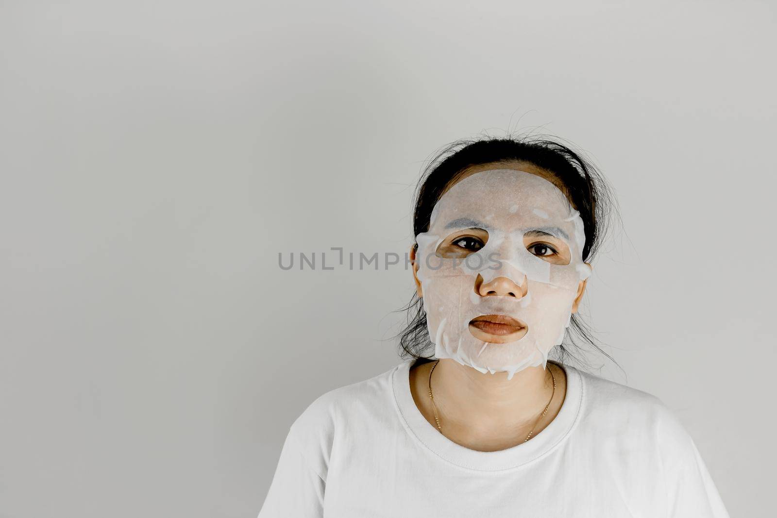 Front view of Asian woman wearing white t-shirt and covering her face with a sheet mask. by wattanaphob