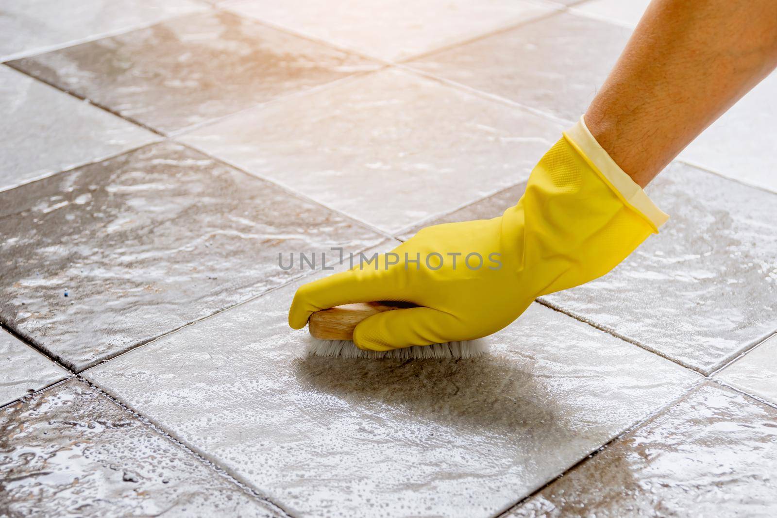 Hands wearing yellow rubber gloves are using a wooden floor scrubber to scrub the tile floor with a floor cleaner.